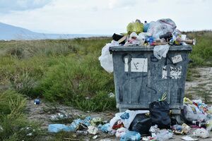 A crowded dumpster stands in nature and overflows.